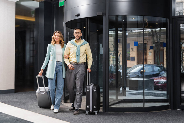 Sorridente coppia interrazziale a piedi con valigie vicino all'hotel  - Foto, immagini