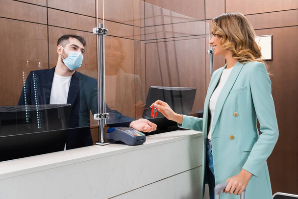 Mujer alegre sosteniendo la llave cerca de la recepcionista en máscara médica en el hotel  - Foto, Imagen