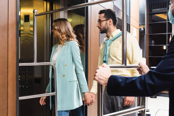 Casal inter-racial em pé perto do elevador e porteiro em máscara médica em primeiro plano desfocado no hotel  - Foto, Imagem