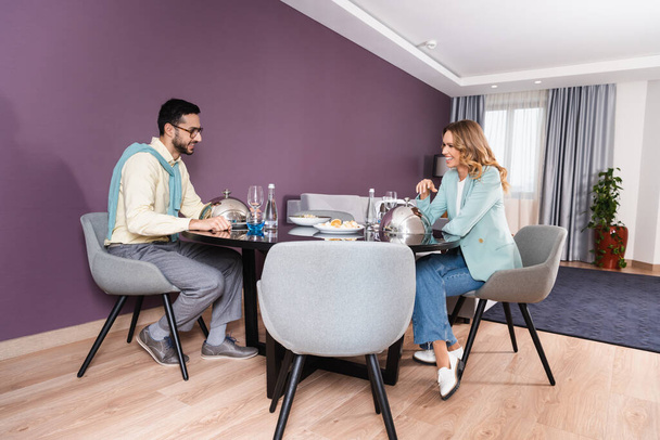 Cheerful interracial couple sitting near dinner in modern hotel room  - Photo, image