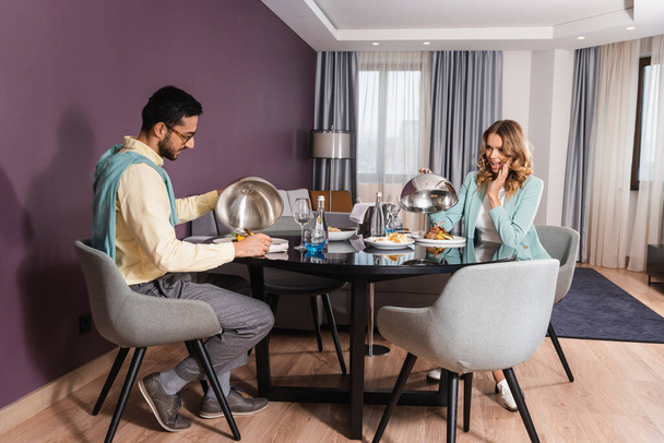Excited woman holding cloche near food and arabian boyfriend in hotel room  - Foto, Imagem