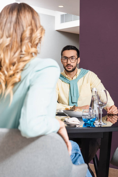 Müslüman bir adam oteldeki yemek masasının yanında bulanık bir ön planda kız arkadaşıyla konuşuyor.  - Fotoğraf, Görsel