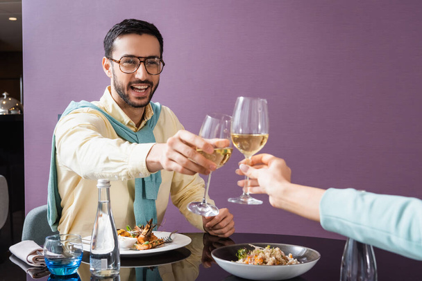 Cheerful arabian man clinking wine with girlfriend near dinner in hotel room  - Fotó, kép