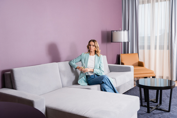 Blonde Frau hält Weinglas auf Sofa im Hotelzimmer  - Foto, Bild