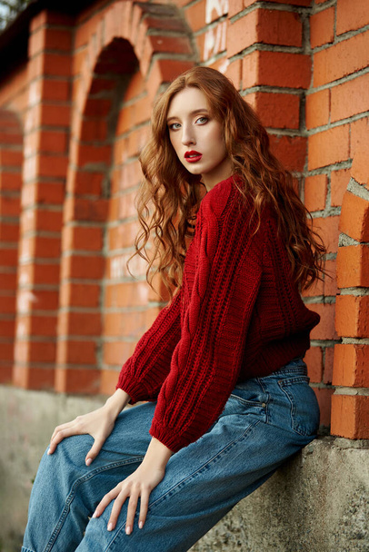 Redhead young woman in a red sweater walks in the park. Autumn beauty portrait of a fashionable Red-haired woman at sunset - Photo, Image