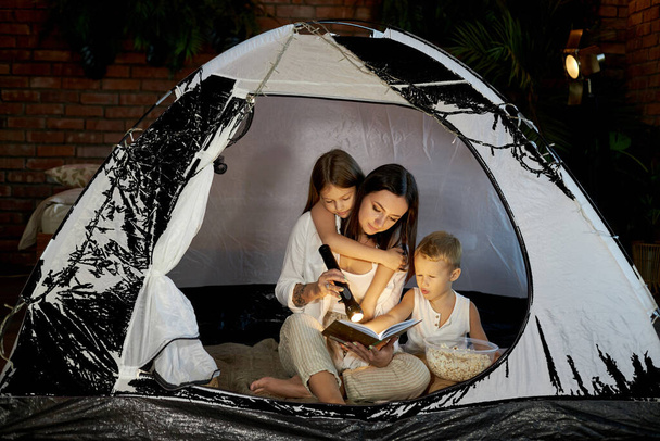 Mom reads children a bedtime story sitting in a tent at home. Mother son and daughter hug and read a book with a flashlight in their hands - Foto, Imagem