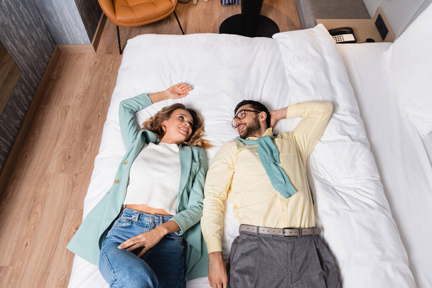 Vista ad alto angolo di allegra coppia interrazziale che si guarda mentre si sdraia sul letto dell'hotel  - Foto, immagini