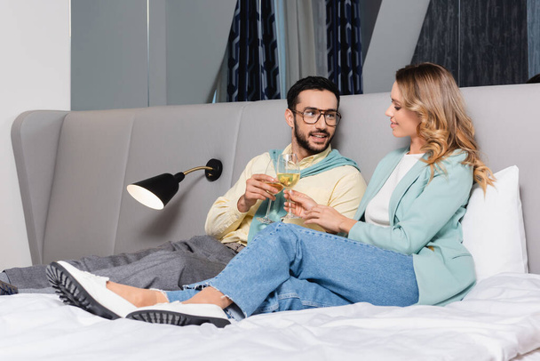 Sonriente pareja interracial brindis con vino en la cama del hotel  - Foto, imagen