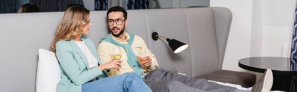 Coppia interrazziale che tiene bicchieri di vino sul letto in camera d'albergo, banner  - Foto, immagini