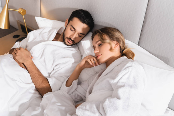 Korkea kulma näkymä rotujenvälinen pari kylpytakit nukkuu hotellihuoneessa  - Valokuva, kuva