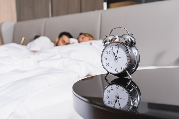 Alarm clock near sleeping couple on blurred background in hotel  - Фото, изображение