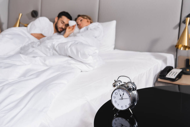 Alarm clock on table near couple sleeping on blurred background in hotel room  - Foto, Imagem