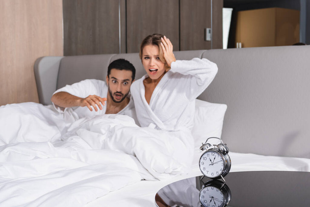 Alarm clock on table near excited interracial couple on hotel bed on blurred background  - Photo, Image