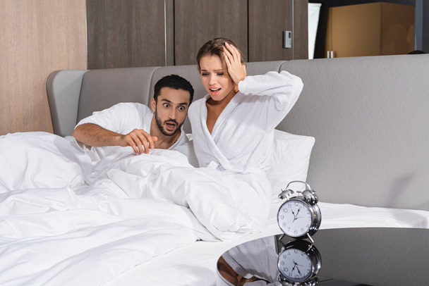 Worried interracial couple on bed near alarm clock in hotel room  - Photo, image