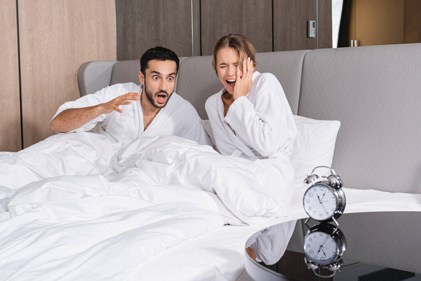 Casal interracial estressado em roupões de banho sentados na cama perto do despertador em primeiro plano desfocado no hotel  - Foto, Imagem