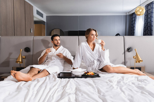 Interracial couple with cups sitting near breakfast on hotel bed  - Fotó, kép