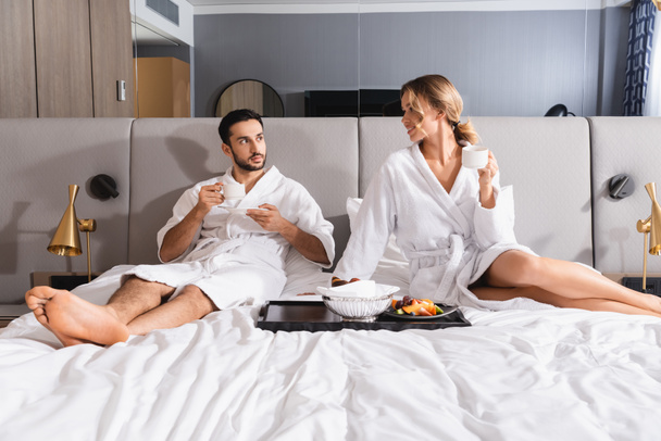 Smiling woman with cup looking at arabian man near breakfast on bed in hotel  - Foto, Imagem