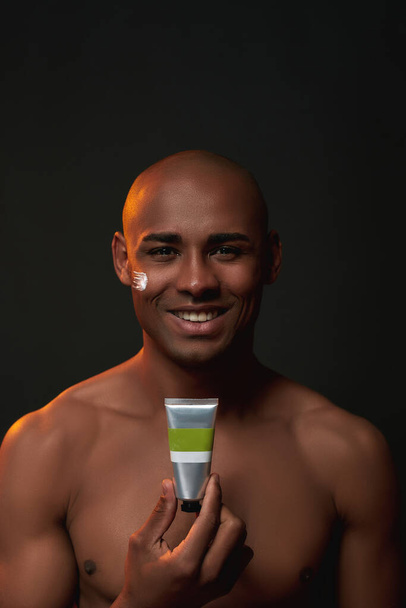 Портрет африканского мужчины без рубашки, улыбающегося в камеру, держащего косметический продукт, позируя со сливками на щеке, изолированном на черном фоне - Фото, изображение