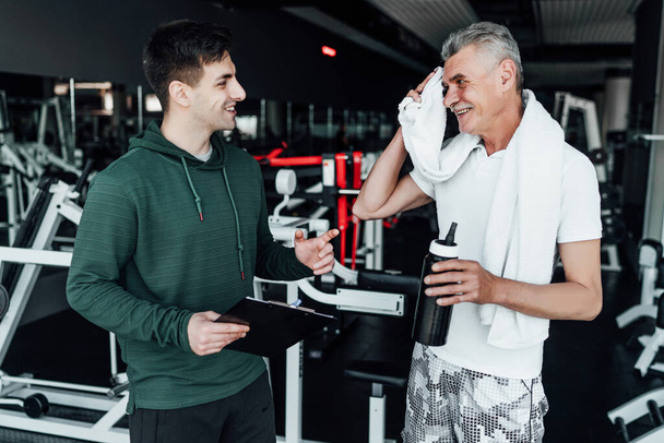 An older man communicates with his coach, they stand in the middle of a modern gym. Sports, health. Rehabilitation specialist, coach. - Fotoğraf, Görsel