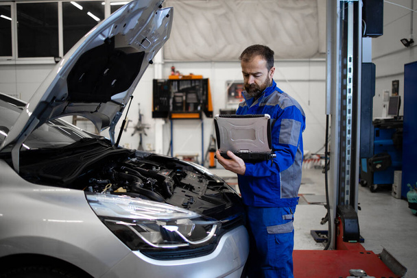 Professional middle aged caucasian car mechanic with laptop computer diagnostic tool standing by vehicle engine area with hood open detecting malfunction. - Photo, Image