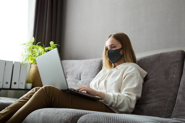 Beautiful young woman in protective medical mask have remote work or online study from home. Cozy home office on couch during coronavirus covid 19 lockdown. freelancer remote work using laptop - 写真・画像