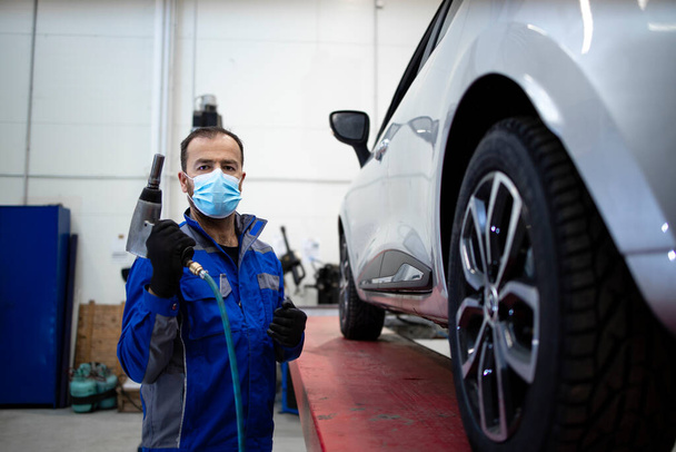 Portrait of professional car mechanic wearing face mask due to corona virus holding pneumatic screw gun and changing wheels of vehicle. Automobile service and maintenance. - Фото, зображення