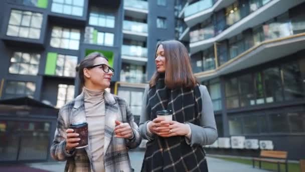Zwei glückliche Frauen spazieren mit Kaffee zum Mitnehmen und unterhalten sich interessiert untereinander im Geschäftsviertel. - Filmmaterial, Video