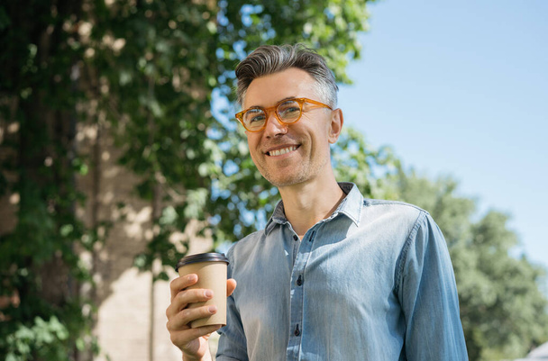 Usmívající se dospělý podnikatel pije kávu venku. Šťastný pohledný muž se stylovými brýlemi, který drží šálek čaje a dívá se do kamery. Koncept přestávky na kávu - Fotografie, Obrázek