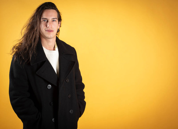 jeune homme aux cheveux longs, avec un visage sérieux en vêtements d'hiver, posant pour la caméra, fond jaune, espace de copie - Photo, image