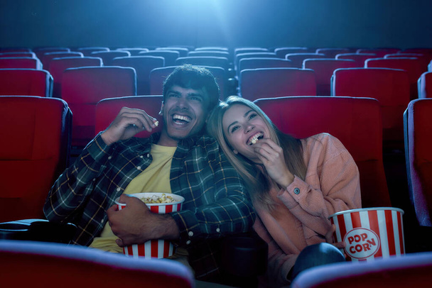 Cheerful young couple, attractive man and woman smiling, sitting at the cinema, watching a movie and eating popcorn - Фото, изображение