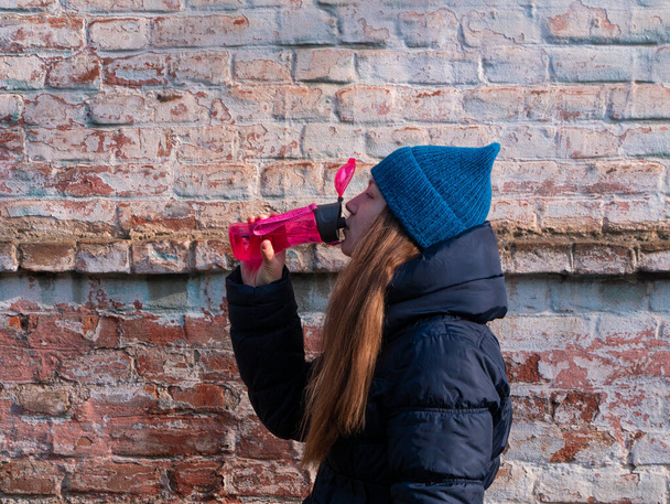 Young woman holds pink reusable bottle of water in hand and drinks on brick wall urban background. Portrait of pretty girl in warm winter clothes on sunny day. Weight loss, active lifestyle body care. - Φωτογραφία, εικόνα