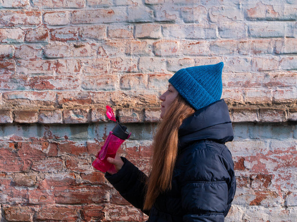 Young woman holds pink reusable bottle of water in hand and drinks on brick wall urban background. Portrait of pretty girl in warm winter clothes on sunny day. Weight loss, active lifestyle body care. - Photo, Image