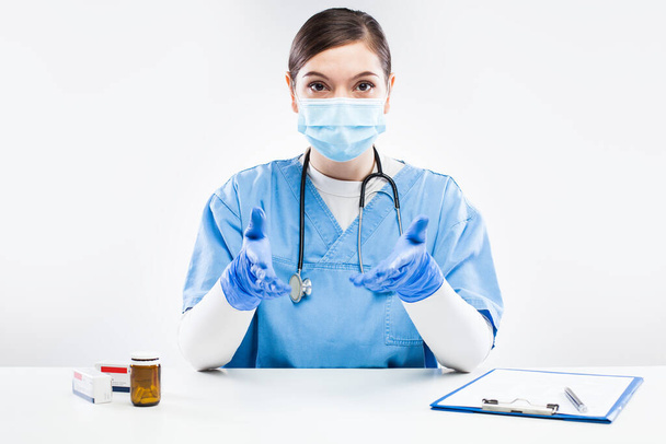 Νεαρή γυναίκα γιατρός του Ηνωμένου Βασιλείου NHS φορώντας προστατευτικό γάντι και μάσκα προσώπου, μιλώντας σε απευθείας σύνδεση με ασθενή, νεαρή γυναίκα GP σε μπλε στολή συμβουλευτικό πρόσωπο εξ αποστάσεως, για τη ζήτηση βοήθεια e-doctor, έννοια τηλεϊατρική - Φωτογραφία, εικόνα