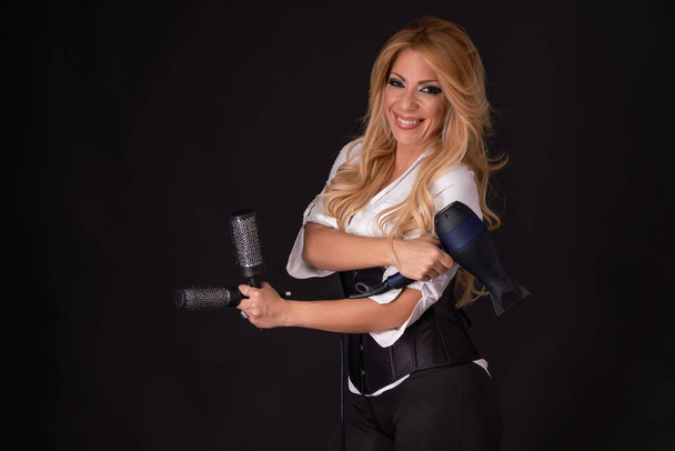 Blonde hairdresser holding a hairdryer and hair brushes while smiling on a black background. - Foto, Bild