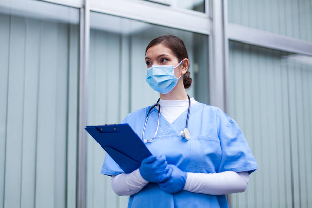 Mulher UK NHS ICU médico trabalhador, mulher médico segurando prancheta vestindo EPI azul proteção esfrega máscara facial, médico de emergência da linha de frente, crise pandêmica COVID-19, triagem de quarentena paciente Coronavirus - Foto, Imagem
