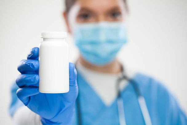 Female GP UK NHS general practitioner doctor,wearing scrubs,protective face mask and gloves,holding white blank bottle of pills,empty no label container suitable for adding custom text,selective focus - Фото, изображение