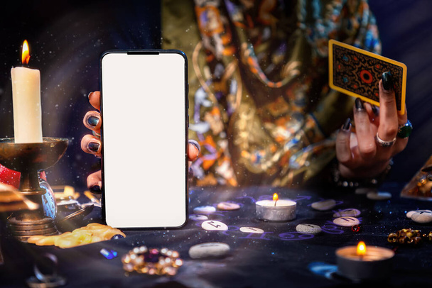 El adivino tiene una tarjeta del Tarot y un smartphone con pantalla blanca. Aura mágica. Prepárate. Primer plano. El concepto de adivinación, magia y esoterismo. - Foto, Imagen