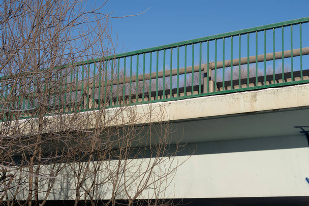 Concrete beam of car bridge, bare branches and green railings against blue sky, background - Photo, Image