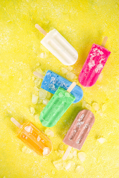 Selection of bright multicolored ice cream popsicle. Various gelato, frozen lollypops - chocolate vanilla blueberry strawberry pistachio orange, with crushed ice on trendy yellow sunny background - Foto, Bild