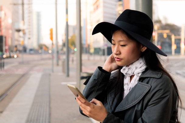 portrait d'une élégante jeune femme asiatique consultant son téléphone portable en attendant à l'arrêt de tramway, concept de transport en commun, technologie et mode de vie urbain - Photo, image