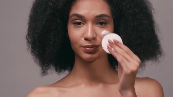 sorridente afro-americana limpeza rosto com leite cosmético isolado em cinza - Filmagem, Vídeo