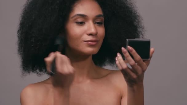 mujer joven afroamericana aplicando polvo facial aislado en gris - Metraje, vídeo