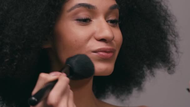 mujer afroamericana aplicando polvo facial con cepillo cosmético aislado en gris - Metraje, vídeo