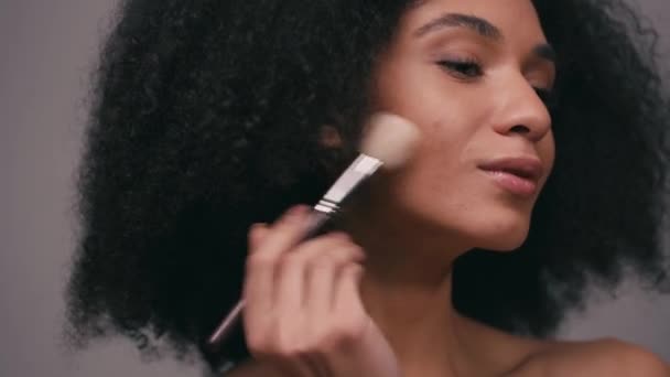 mujer afroamericana aplicando iluminador con cepillo cosmético aislado en gris - Metraje, vídeo