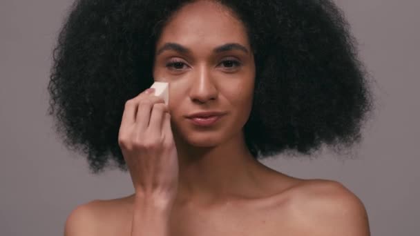 african american woman applying makeup foundation with sponge isolated on grey - Záběry, video