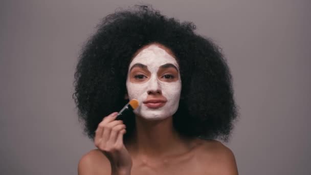 sonriente mujer afroamericana aplicando máscara de arcilla aislada en gris - Imágenes, Vídeo