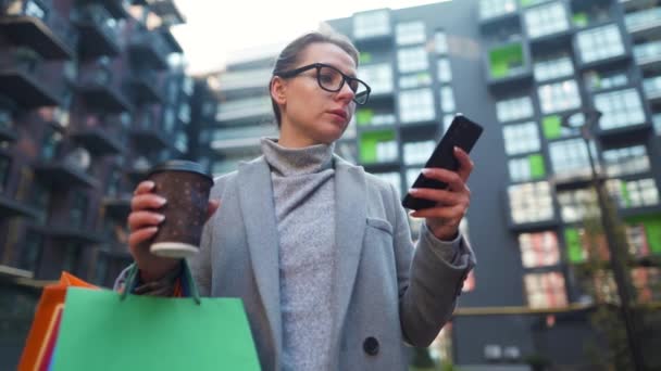 Женщина стоит в деловом районе, держит на вынос кофе, сумки и использует смартфон. Концепция городского образа жизни и цифровых технологий - Кадры, видео