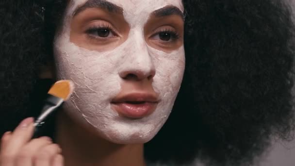 Afrykańska amerykanka nakładająca glinianą maskę ze szczotką kosmetyczną wyizolowaną na szaro - Materiał filmowy, wideo