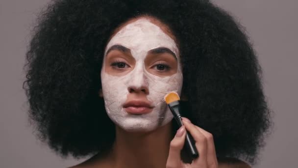 mujer afroamericana mirando a la cámara y aplicando máscara de arcilla aislada en gris - Imágenes, Vídeo