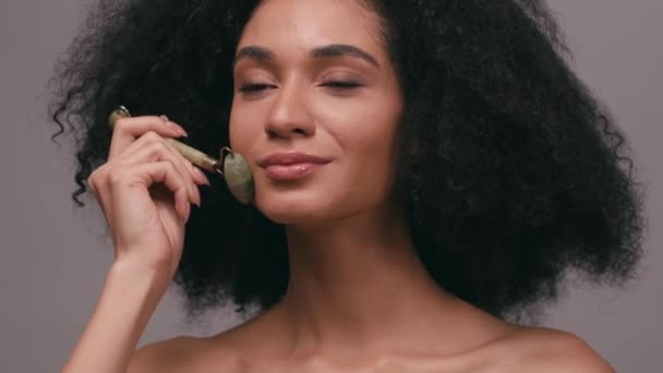 Африканська американка, яка користується нефритовим роликом на обличчі і шиї ізольованих на сірому - Кадри, відео
